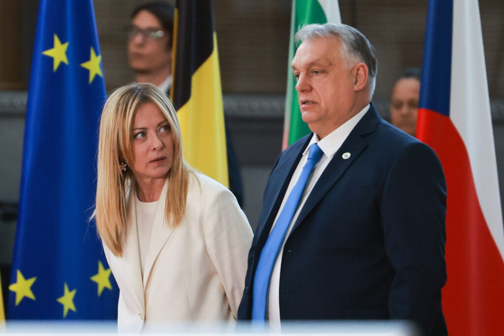 Italiens Ministerpräsidentin Giorgia Meloni blickt seitlich auf ihren neben ihr stehenden ungarischen Amtskollegen Viktor Orbán. Im Hintergrund die Fahnen mehrerer Nationen. Das Foto wurde während einem formellen Treffen des Rats der Europäischen Union im März 2026 gemacht.