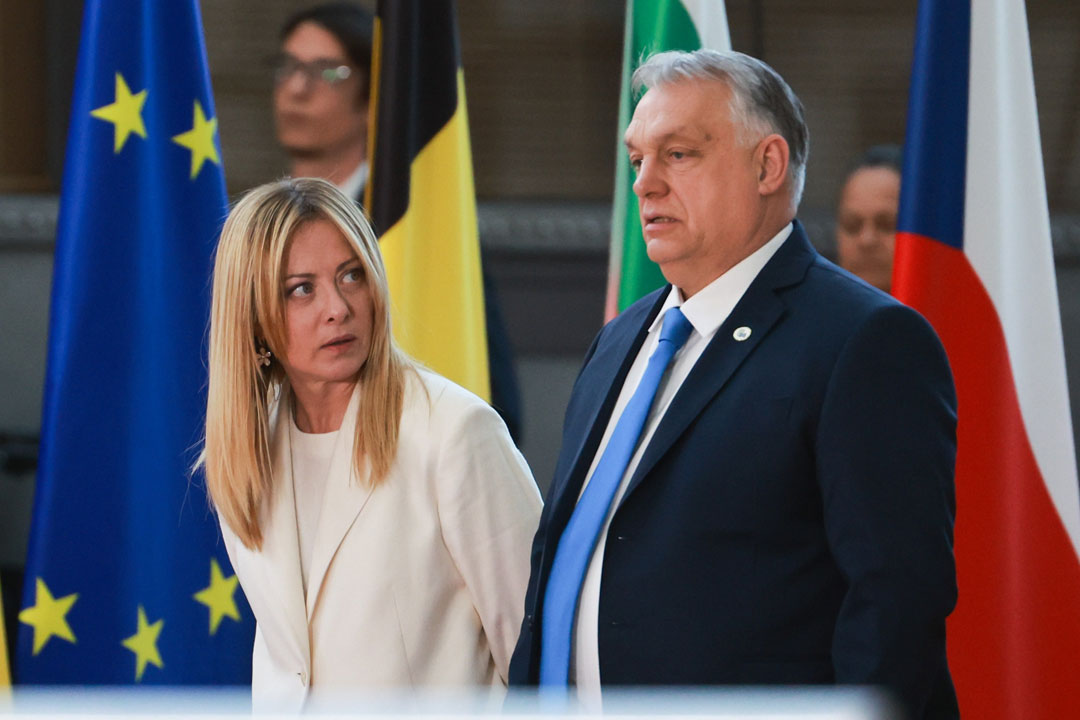 Italiens Ministerpräsidentin Giorgia Meloni blickt seitlich auf ihren neben ihr stehenden ungarischen Amtskollegen Viktor Orbán. Im Hintergrund die Fahnen mehrerer Nationen. Das Foto wurde während einem formellen Treffen des Rats der Europäischen Union im März 2026 gemacht.