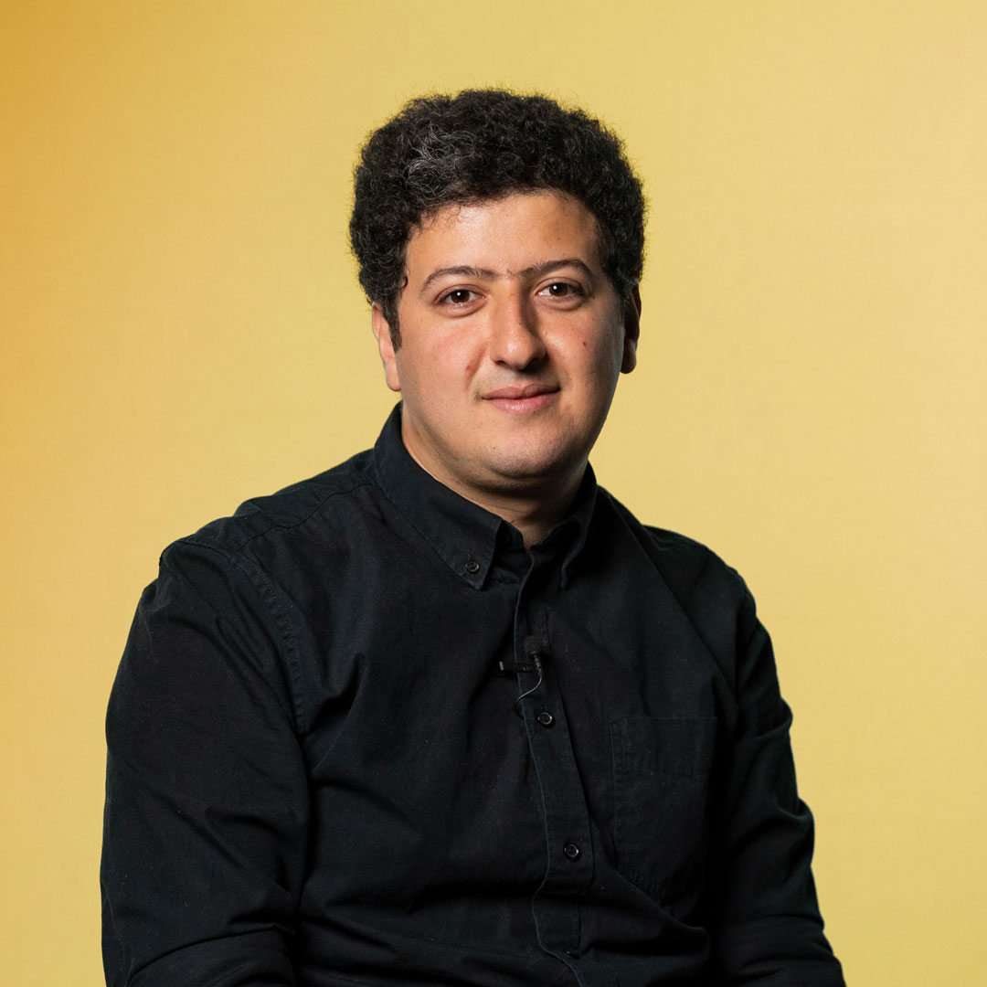 Portrait d’un homme sur fonds jaune, cheveux foncés et souriant, le visage tourné vers l’appareil photographique.