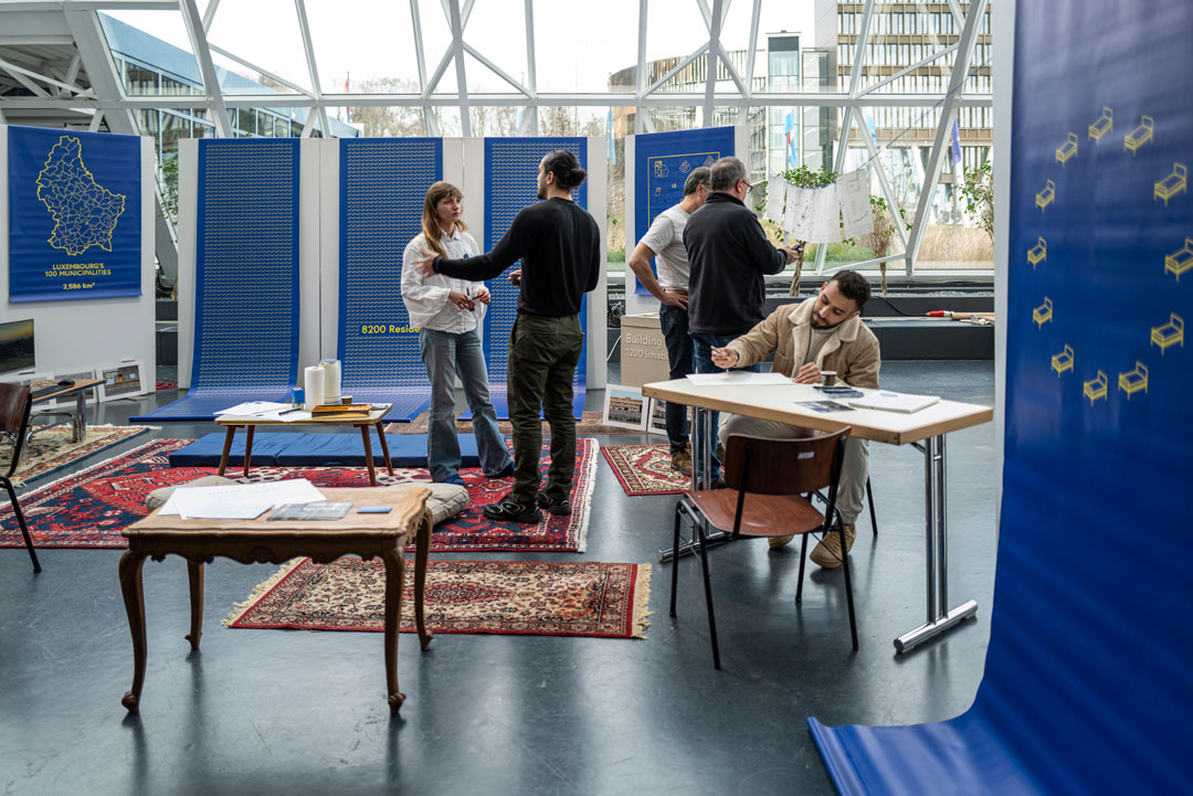 Cinq personnes dans un bâtiment éclairé, des fenêtres au fond, des tapis sur le sol. Ils participent à un atelier, les personnnes parlent entre elles et une est assise à un table, en dessinant ou écrivant sur un papier.