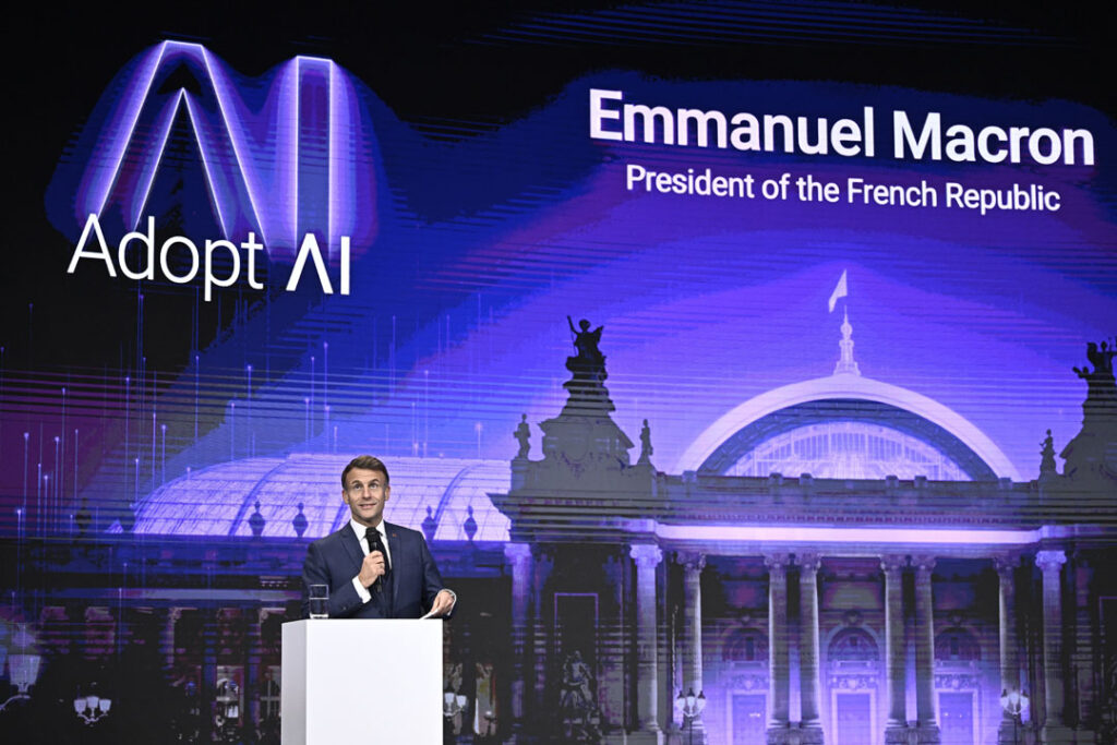 Der französische Präsident Emmanuel Macron steht auf einer in dunkelblau gehaltenen Bühne und hält eine Rede; im Hintergrund ist der Schriftzug „Adopt AI“, so der Name der Konferenz, zu sehen.