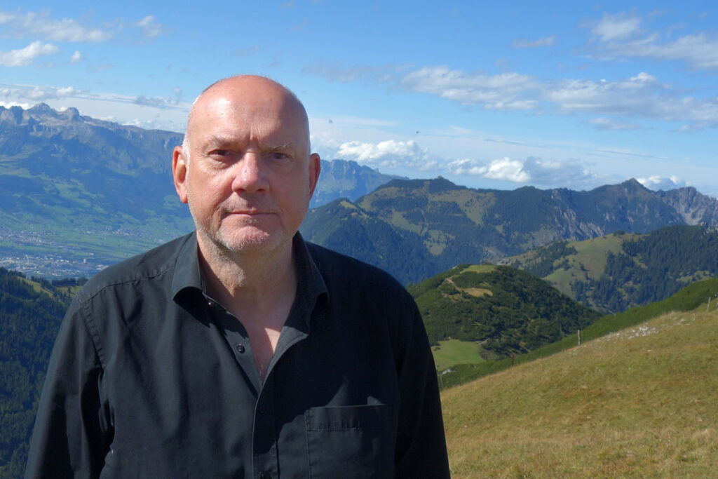 Der Autor Guy Helminger mit ernstem Gesichtsausdruck vor einer Berglandschaft.