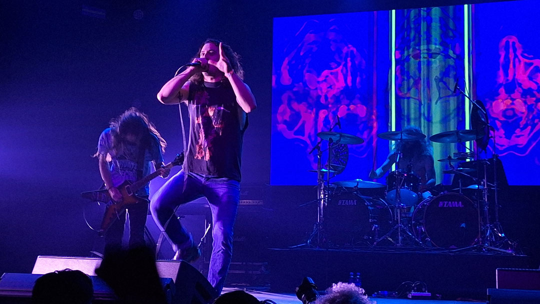 Auftritt der Metal-Band Inter Arma. Der Sänger stützt sich mit einem Fuß auf den Backline-Monitor. Er hält mit der einen Hand das Mikrofon und gestikuliert mit der anderen. Neben und hinter ihm sind ein Gitarrist und der Schlagzeuger zu sehen.