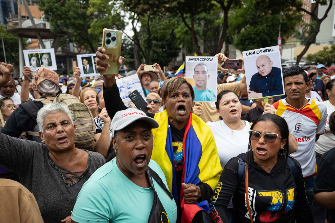 Das Bild zeigt eine Gruppe von Demonstrierenden, die Porträts mit politischen Gefangenen und venezolanische Nationalfahnen mit sich führen. 