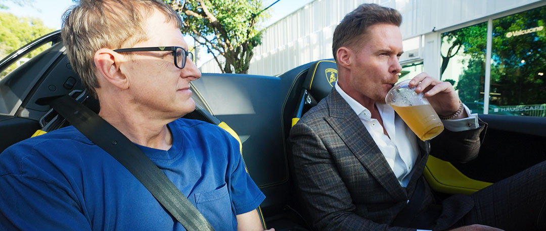 Louis Theroux und Justin Waller sitzen im Cabrio-Porsche von letzterem. Waller hat ein Getränk in der Hand und trägt einen teuren Anzug, Theroux schaut skeptisch.