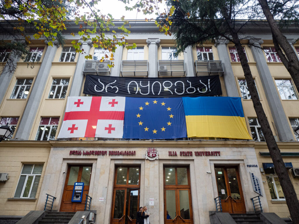 Fassade der Ilia-Universität im Zentrum von Tiflis; am unteren Bildrand zwei Studentinnen. Über dem Eingangsbereich mit vier großen Flügeltüren hängen nebeneinander die Fahnen Georgiens, der Europäischen Union und der Ukraine.