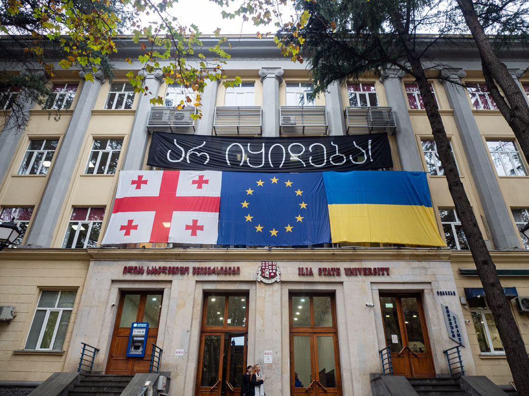 Fassade der Ilia-Universität im Zentrum von Tiflis; am unteren Bildrand zwei Studentinnen. Über dem Eingangsbereich mit vier großen Flügeltüren hängen nebeneinander die Fahnen Georgiens, der Europäischen Union und der Ukraine.