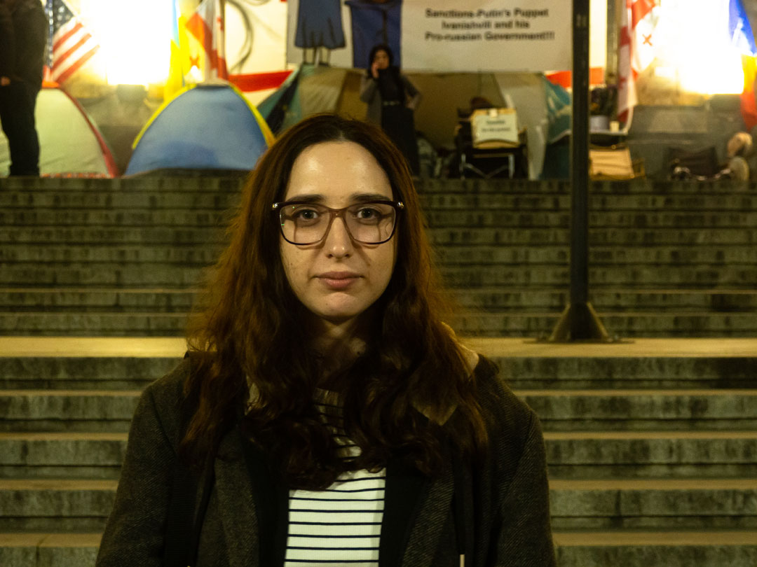 Frontalporträt der Jurastudentin Lizi Basilaia, vor einem Treppenaufgang; im Bildhintergrund sind vor dem Gebäude aufgerichtete Zelte zu sehen, was nahelegt, dass das Bild während einer Protestaktion aufgenommen wurde. Verschwommen ist ein Plakat mit einer Parole zu erkennen.