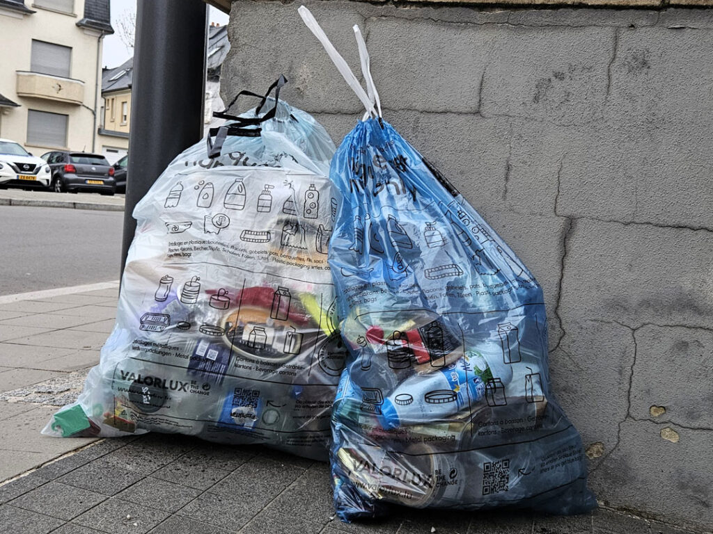 Zwei Plastiksäcke liegen auf Straße, sie sind mit Plastikmüll wie Becher gefüllt.