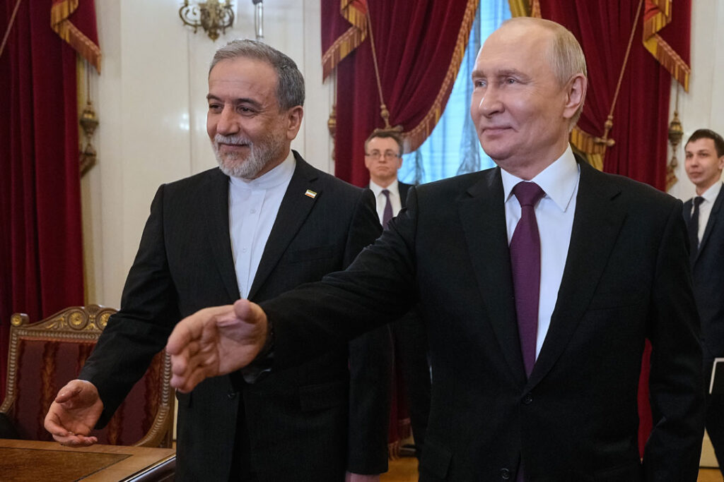 Das Bild zeigt den russischen Präsidenten Wladimir Putin und den iranischen Außenminister Abbas Araghchi nebeneinanderstehend. Beide strecken einer oder mehreren Personen die Hand zum Handschlag entgegen. Die betreffenden Personen sieht man nicht. Im Bildhintergrund ein festlich geschmückter Saal und Männer in Anzügen, was den offiziellen Charakter des Rahmens unterstreicht.