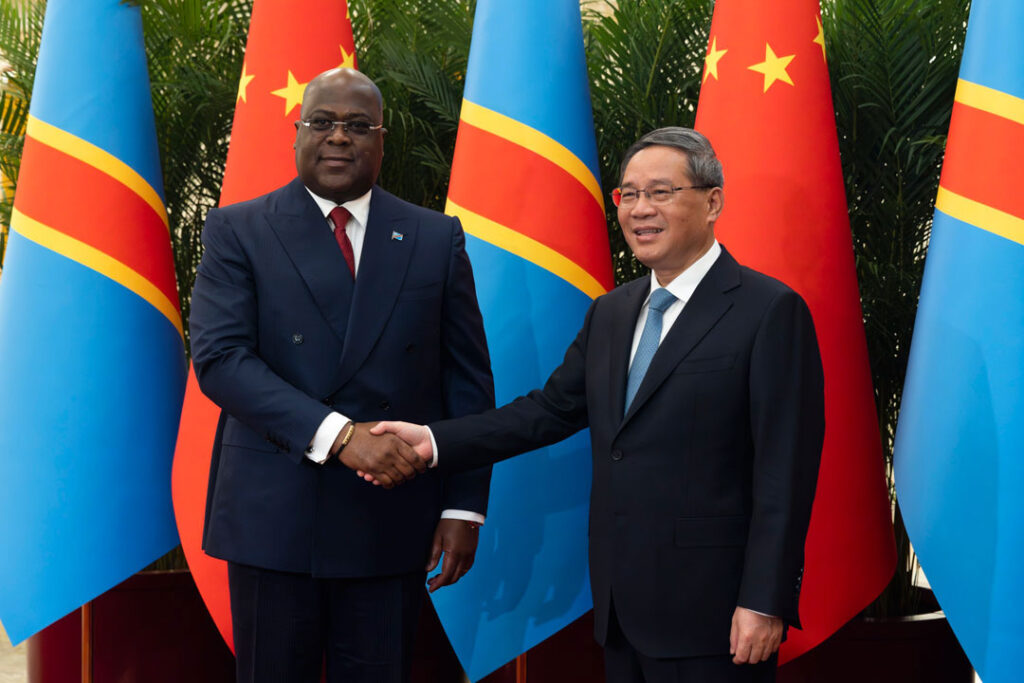 Das Bild zeigt den kongolesischen Präsidenten Félix Tshisekedi und den chinesischen Ministerpräsidenten Li Qiang, die sich die Hand schütteln. Im Bildhintergrund sind mehrere, sich abwechselnde Fahnen der Demokratischen Republik Kongo und der Volksrepublik China zu sehen.
