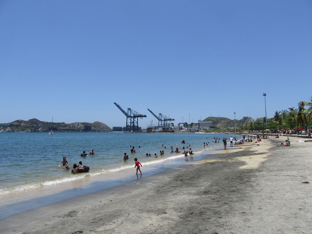 Die Stadt Santa Marta am Meer, ein Strand im Vordergrund, im Hintergrund ein Hafen mit Kohle, Kinder spielen auf dem Strand.