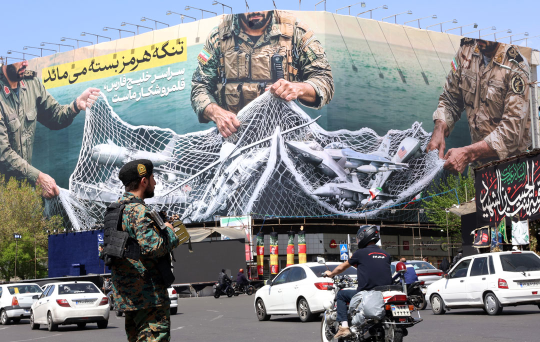 Das Bild zeigt eine Straßenszene am zentral gelegenen Enghelb-Platz in der iranischen Hauptstadt Teheran. Im Bildvordergrund steht ein Soldat mit geschultertem Gewehr, vor ihm passieren einige Autos und ein Motorrad. Der Bildhintergrund wird durch ein großes Gebäude ausgefüllt, dessen Fassade vollständig in ein Transparent gehüllt ist. Darauf sind verschiedene Militärs zu sehen, die ein Fischernetz einholen, in dem US-amerikanisches Militärgerät, vor allem Flugzeuge und ein Kriegsschiff, gefangen sind.