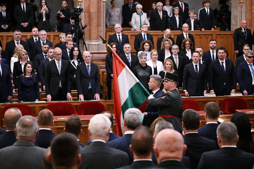Unser Bild zeigt das ungarische Parlament bei seiner konstituierenden Sitzung. Im Bildvordergrund, von hinten, die Abgeordneten, im Bildhintergrund, frontal, die Regierungsbank. In der Bildmitte schreitet der neue Ministerpräsident Péter Magyar von rechts nach links durch den Saal; er wird begleitet von einem uniformierten Träger der ungarischen Fahne.