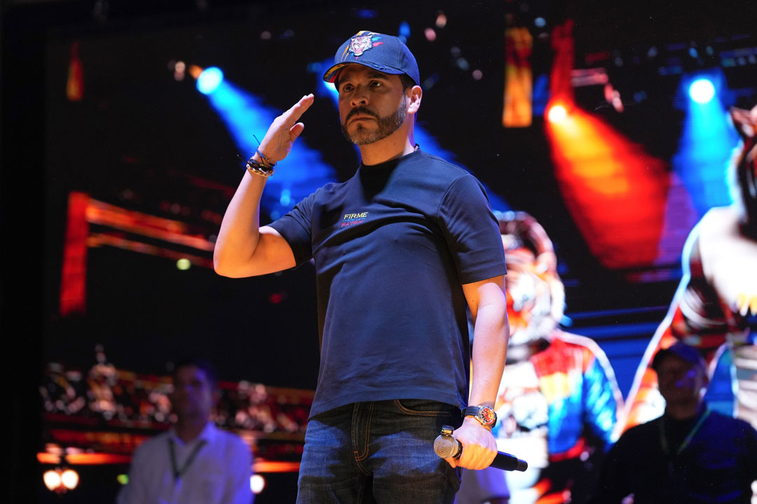 Das Bild zeigt den rechtsextremen kolumbianischen Präsidentschaftskandidaten Abelardo de la Espriella auf einer Bühne, die wie bei einem Popkonzert ausgeleuchtet ist. Er ist leger gekleidet, trägt eine Baseballmütze und führt die rechte Hand zum militärischen Gruß an den Kopf, während er in der linken lässig ein Mikro hält.