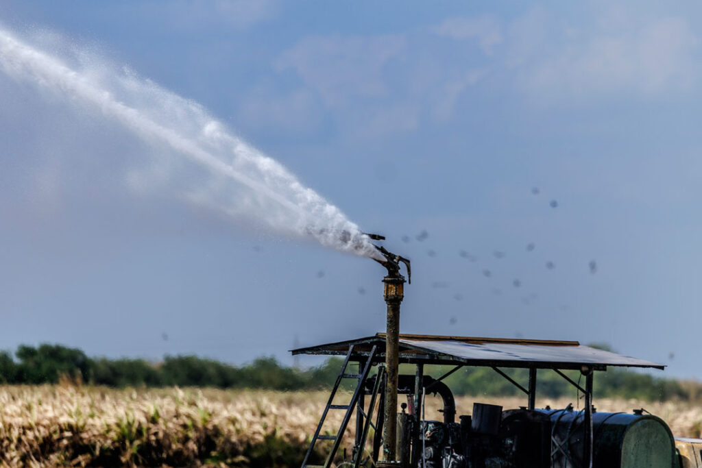 Foto einer Bewässerungsanlage in der Landwirtschaft. Zu sehen ist ein Tank sowie eine Vorrichtung, aus der ein Wasserstrahl spritzt. Im Hintergrund ist ist ein Feld zu erkennen.