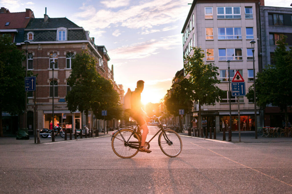 Ein Fahrradfahrer steht auf einer Kreuzung in einer Stadt. Abgesehen von ihm ist die Szene menschenleer. Er schaut in Richtung untergehende Sonne.