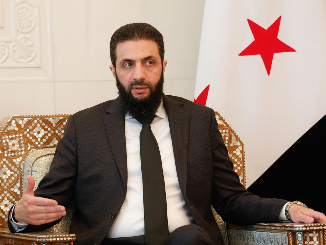 Der syrische Übergangspräsident Ahmed al-Sharaa in einem schlicht geschmückten Sessel sitzend, im Hintergrund ein Ausschnitt der syrischen Fahne.
