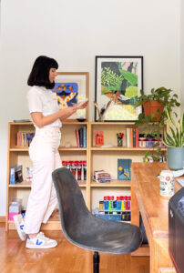 L’artiste Ana Gaman, debout dans son atelier, devant une table et un mur décoré avec des affiches et des illustrations.