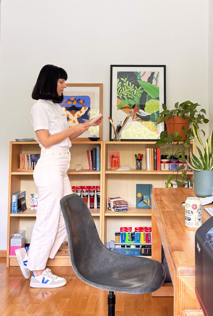 L’artiste Ana Gaman, debout dans son atelier, devant une table et un mur décoré avec des affiches et des illustrations.