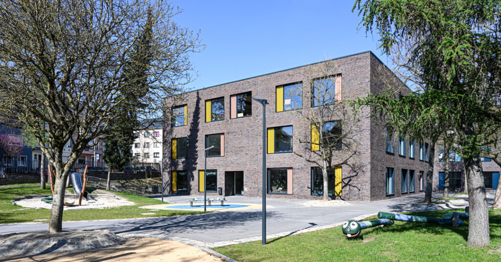Eine Grundschule in Luxemburg-Hollerich. Der moderne Backsteinbau hat große Fenster mit gelben und orangen Vorhängen. Vor dem Gebäude stehen neben Bäumen auch Spielgeräte für Kinder.