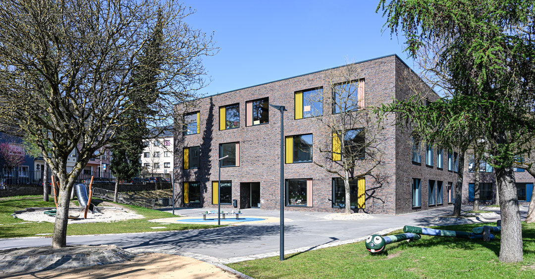 Eine Grundschule in Luxemburg-Hollerich. Der moderne Backsteinbau hat große Fenster mit gelben und orangen Vorhängen. Vor dem Gebäude stehen neben Bäumen auch Spielgeräte für Kinder.