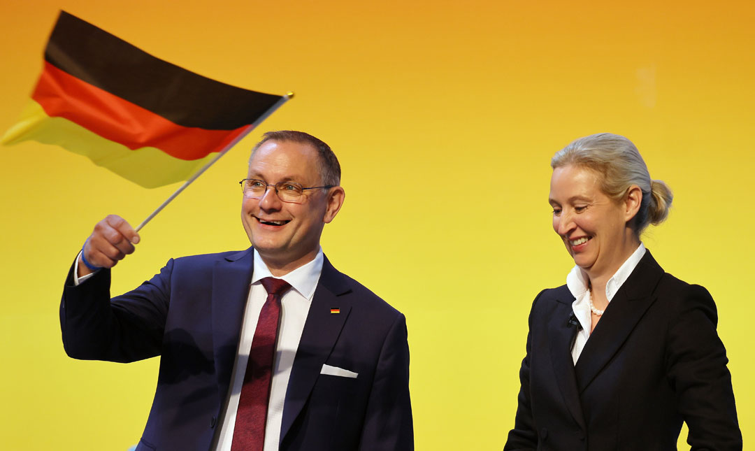 Die Co-Vorsitzenden der AfD, Alice Weidel (rechts) und Tino Chrupalla (links), stehen vor einem gelben Hintergrund lachend nebeneinander, Weidel blickt nach unten, Chrupalla schwenkt eine kleine Deutschlandfahne und blickt ins Publikum.