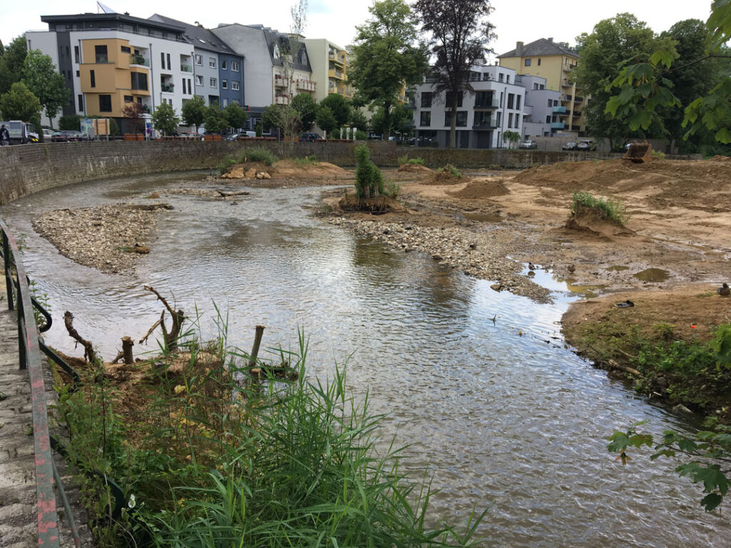 Ein flacher Bach läuft entlang einer Mauer und zwischen Sandhügeln. Im Hintergrund sind einige Familienhäuser und Bäume zu sehen.
