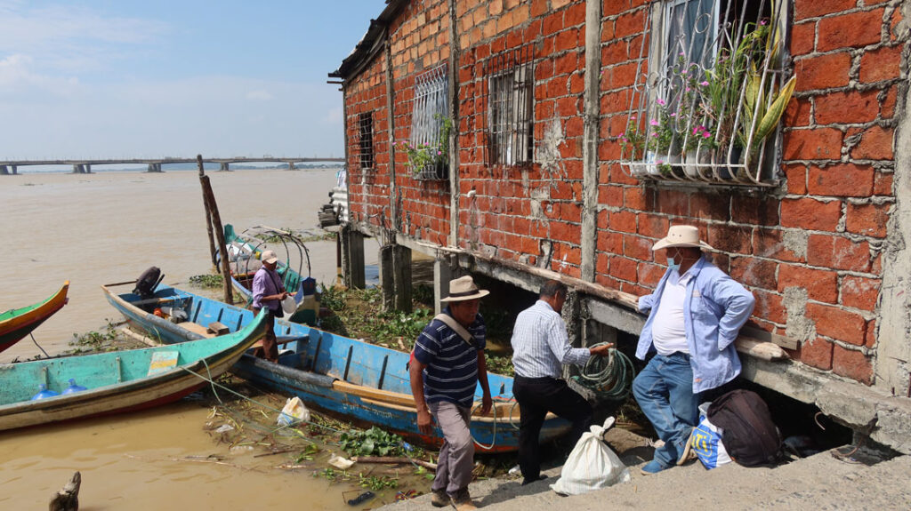 Eine Bootsanlegestelle mit einem Backsteingebäude rechts, das in den Fluss hineinragt. Zwei Langboote sind zu sehen und mehrere Personen; eine hantiert mit einem Tau, eine Mann lehnt an der Hauswand und wartet. Im Hintergrund der Fluss und eine Brücke.