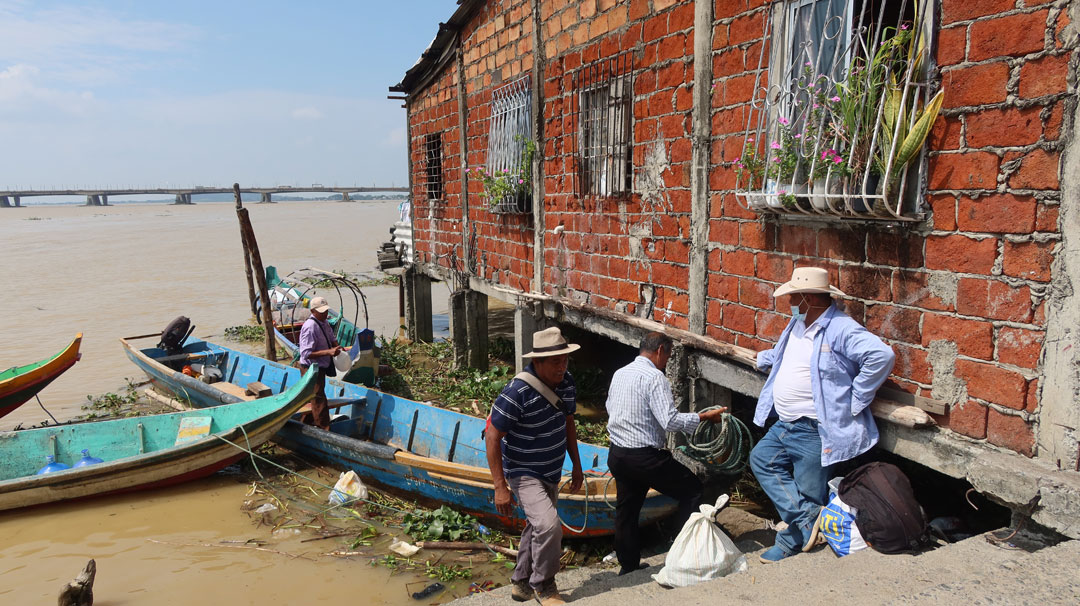 Eine Bootsanlegestelle mit einem Backsteingebäude rechts, das in den Fluss hineinragt. Zwei Langboote sind zu sehen und mehrere Personen; eine hantiert mit einem Tau, eine Mann lehnt an der Hauswand und wartet. Im Hintergrund der Fluss und eine Brücke.