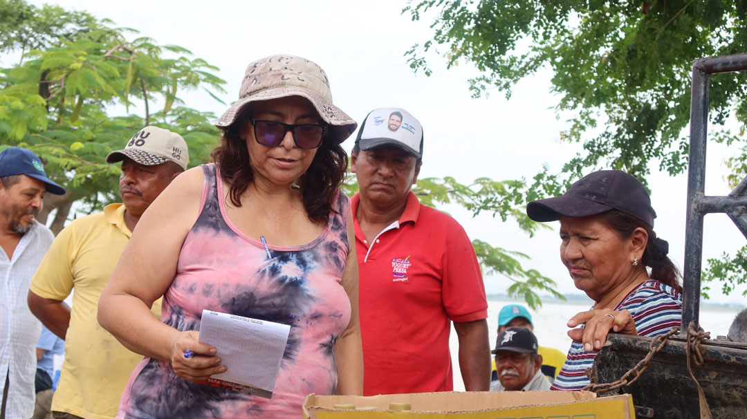 Raquel Silva mit Sonnenhut und Sonnenbrille, einen Schreibblock in der rechten Hand. Sie kontrolliert die Ladung auf einem Pick-Up-Truck. Ein Mann und eine Frau sehen ihr dabei zu.