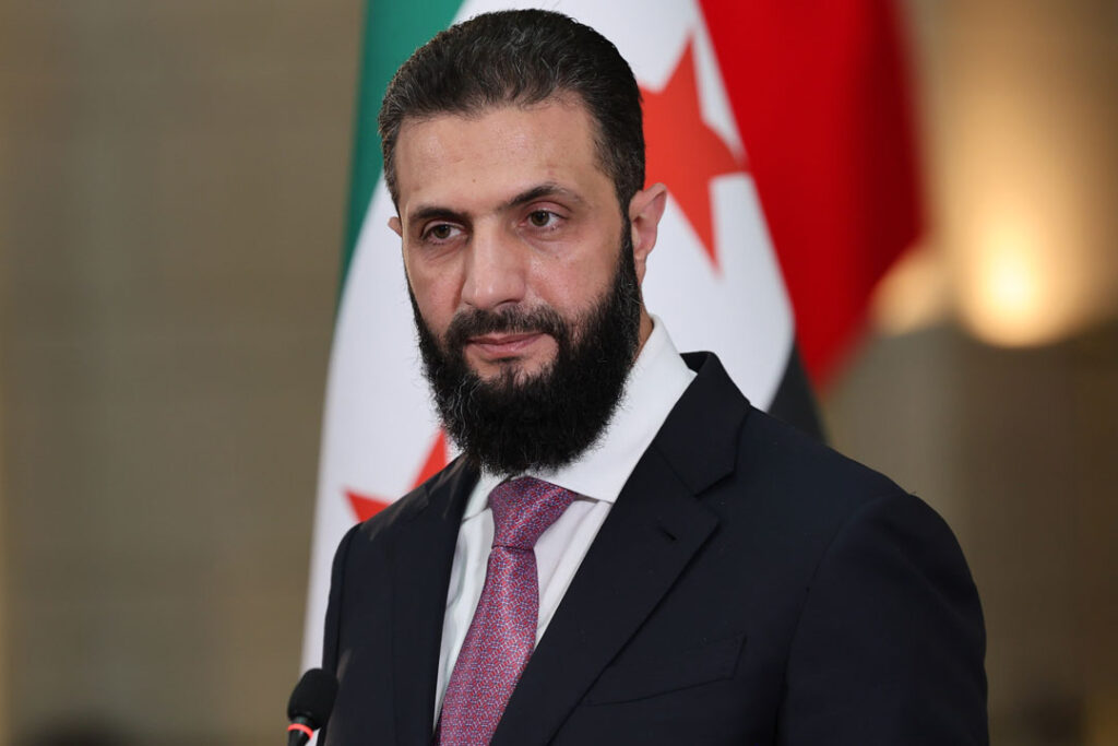 Syriens Präsident Ahmed al-Sharaa in Anzug und Krawatte, im Hintergrund schemenhaft die syrische Fahne.