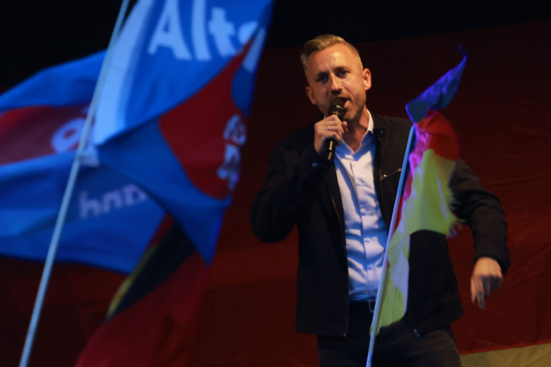 Der AfD-Politiker Ulrich Sigmund mit dem Mikrofon in der Hand auf einer Rednerbühne, im Vordergrund unscharf AfD- und Deutschlandfahnen.