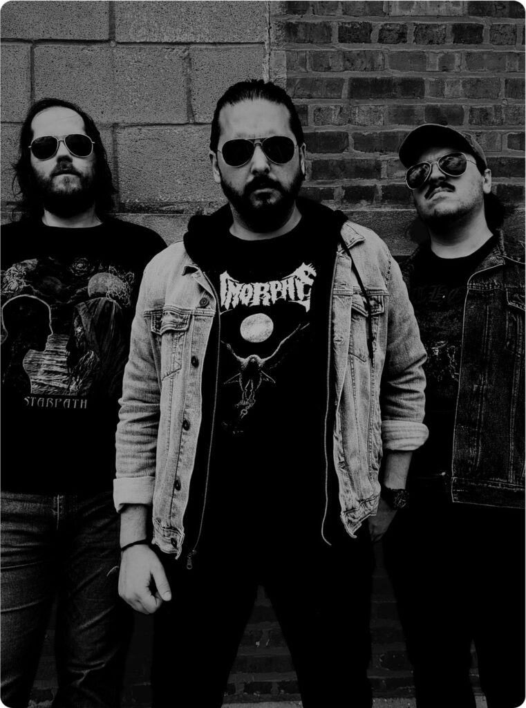 Schwarz-weißes Foto der Band Fer de Lance vor dem Hintergrund einer Hauswand. Die Musiker tragen Bandshirts und Sonnenbrillen.