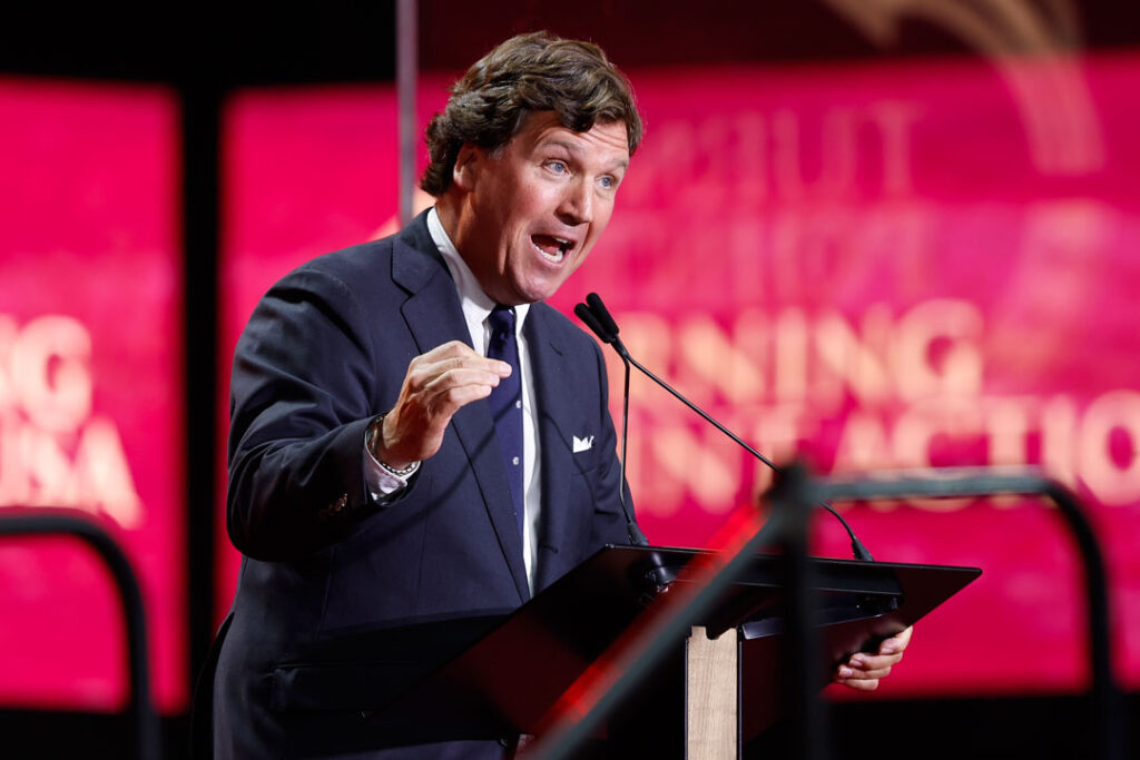 Tucker Carlson, mit einer Hand gestikulierend, die andere am Rednerpult.