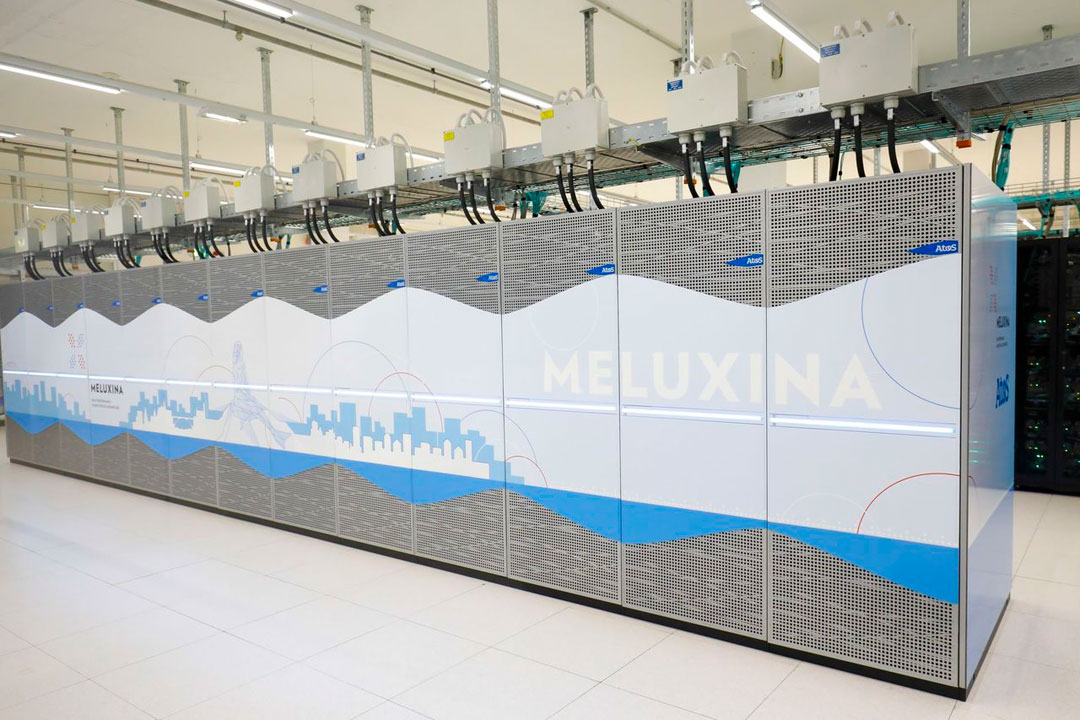 Foto des Supercomputers Meluxina. In einem Rechenzentrum stehen auf weiß gefliestem Boden Schränke, in denen Computer sind. Zu sehen ist aber lediglich die Stromversorgung, die auf einer Metallschiene von der Decke kommt. Die Computerschränke sind mit mit Löchern versehenen Metalltüren verschlossen. Auf diesen Türen ist eine stilisierte Skyline sowie der Name Meluxina aufgedruckt.