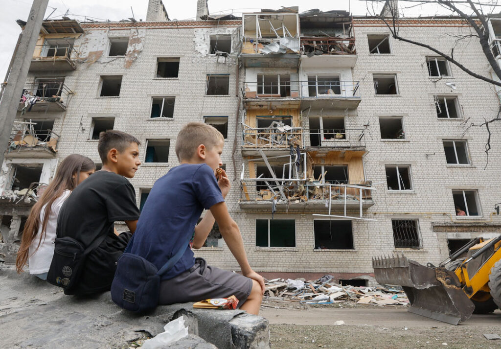 Drei Kinder sitzen auf einer Mauer vor einem mehrstöckigen Wohnhaus, das vorne schwer beschädigt ist; mehrere Balkone wurden abgerissen und es sind keine Fensterscheiben mehr zu sehen.
