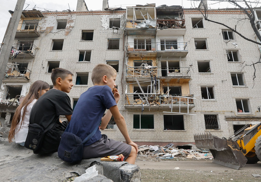 Drei Kinder sitzen auf einer Mauer vor einem mehrstöckigen Wohnhaus, das vorne schwer beschädigt ist; mehrere Balkone wurden abgerissen und es sind keine Fensterscheiben mehr zu sehen.