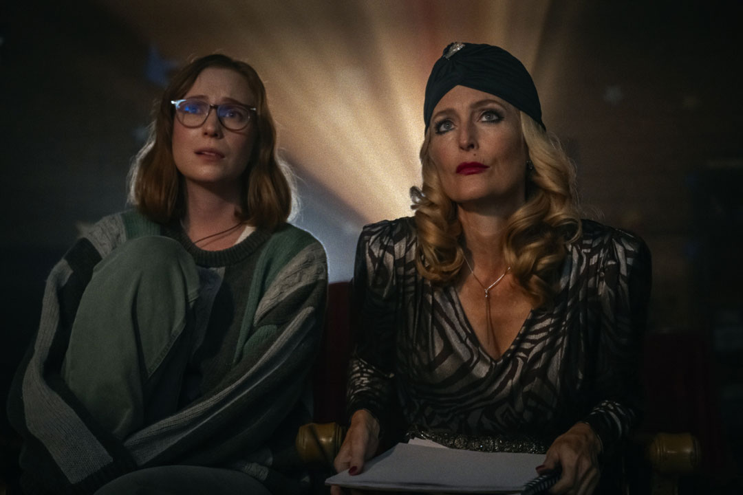 Zwei Frauen sitzen in einem Kino, hinter ihnen leuchtet ein Projektor. Die junge Frau links, locker gekleidet schaut etwas verzweifelt, während die ältere Frau rechts von ihr, gekleidet wie eine Filmdiva mit Turban, konzentriert auf die Leinwand sieht.