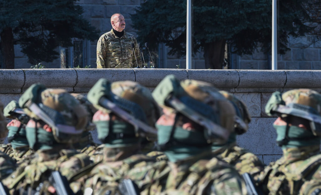 Im Vordergrund sind von der Brust aufwärts fünf marschierende, vermummte Soldaten zu sehen, ihren Blick auf den aserbaidschanischen Präsidenten gerichtet, der im Bildhintergrund in Militärkhaki auf einem Podium steht.
