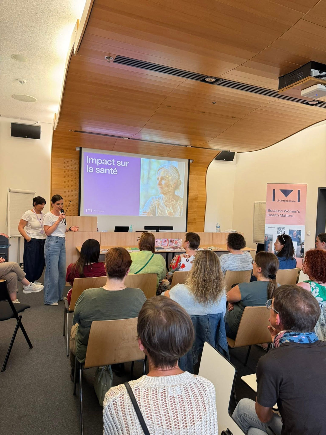 Plusieurs femmes sont assises devant l’image projetée du buste d’une femme dont l’intérieur du corps apparaît en lumineux. Devant l’image, une femme parle dans un micro et une autre se tient derrière elle.