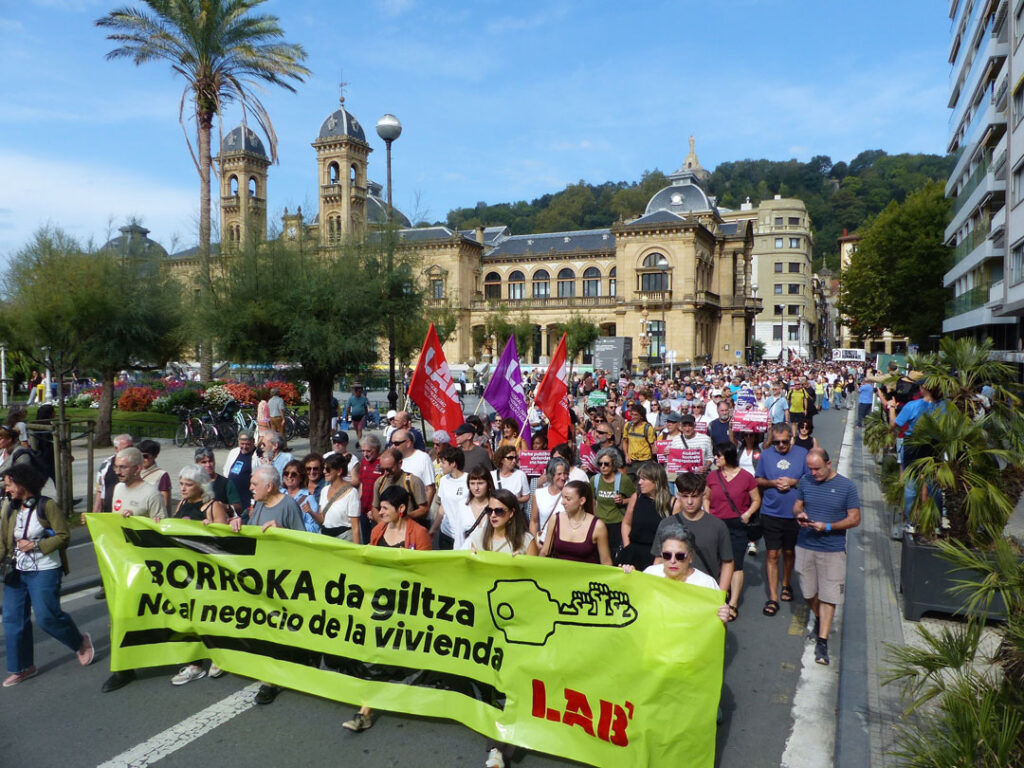 Es sind zahlreiche Teilnehmer*innen einer Demonstration zu sehen. Im Bildvordergrund eine Reihe von Personen, die ein grünes Plastiktransparent mit schwarzer und roter Aufschrift tragen. Im Hintergrund ist das Rathaus von San Sebastián zu sehen.