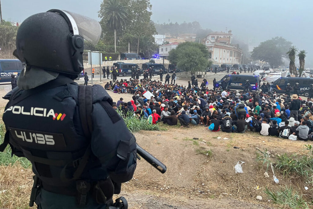 Im Bildvordergrund ist ein Polizist in Kampfmontur und mit Schlagstock zu sehen, in einiger Entfernung von ihm kauern Dutzende Menschen auf dem Boden. Die Szenerie ist von Mannschaftswägen der Polizei gesäumt.