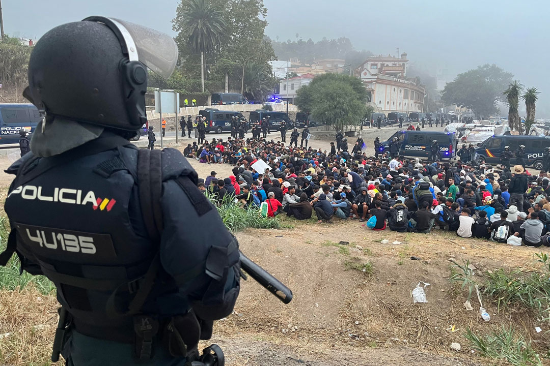 Im Bildvordergrund ist ein Polizist in Kampfmontur und mit Schlagstock zu sehen, in einiger Entfernung von ihm kauern Dutzende Menschen auf dem Boden. Die Szenerie ist von Mannschaftswägen der Polizei gesäumt.