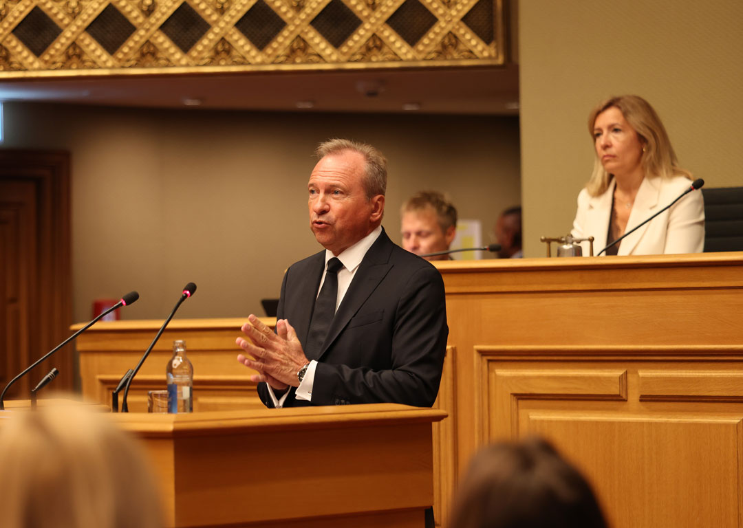 Foto von Gilles Roth im Parlament. Der Finanzminister steht am Rednerpult der Chamber. Er trägt einen schwarzen Anzug und eine dunkle Kravatte, hat seine Hände gefaltet und hat einen nachdenklichen Blick.