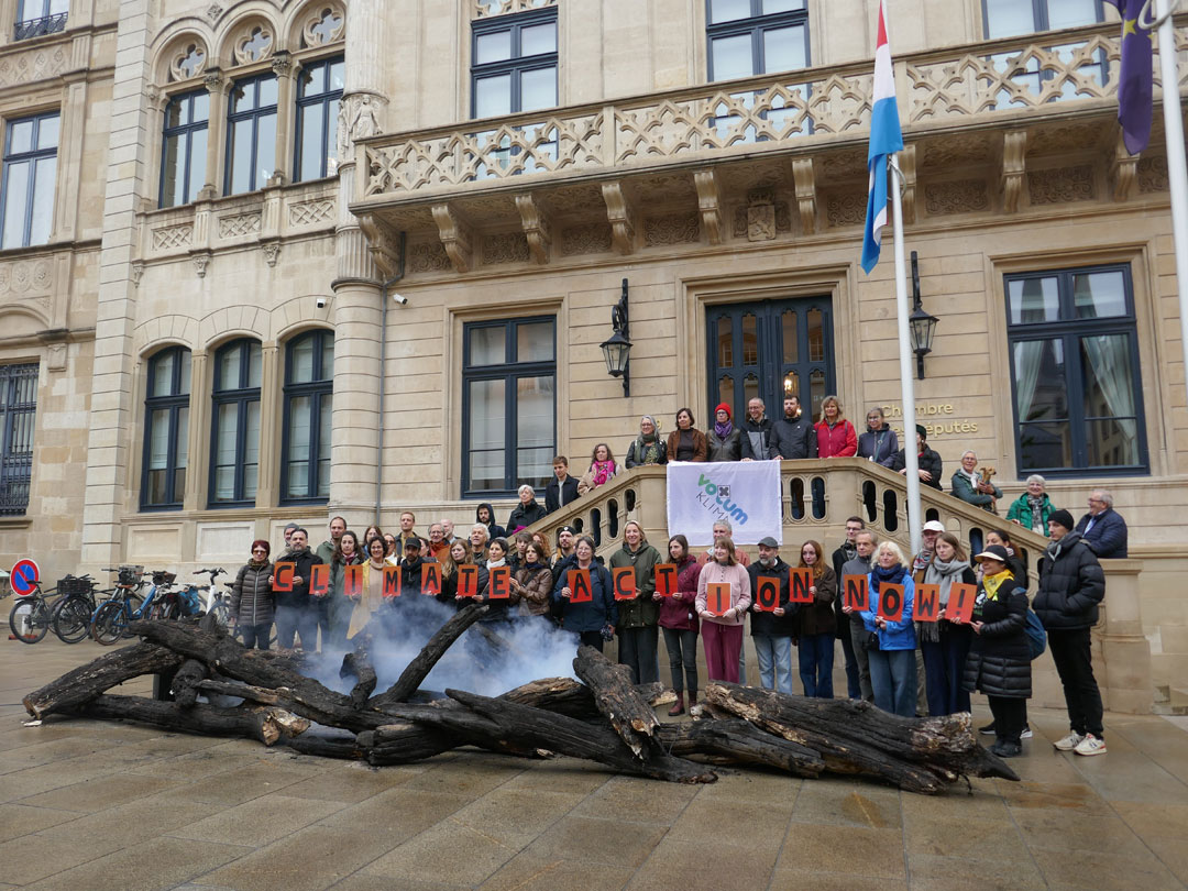 Eine Gruppe von rund fünfzig Menschen stehen vor dem Parlament in Luxemburg-Stadt, hinter verkohlten Baumstämmen aus denen Rauch emporsteigt. Sie halten je einen Buchstaben in der Hand, die zusammen „Climate Action Now!“ ergeben.