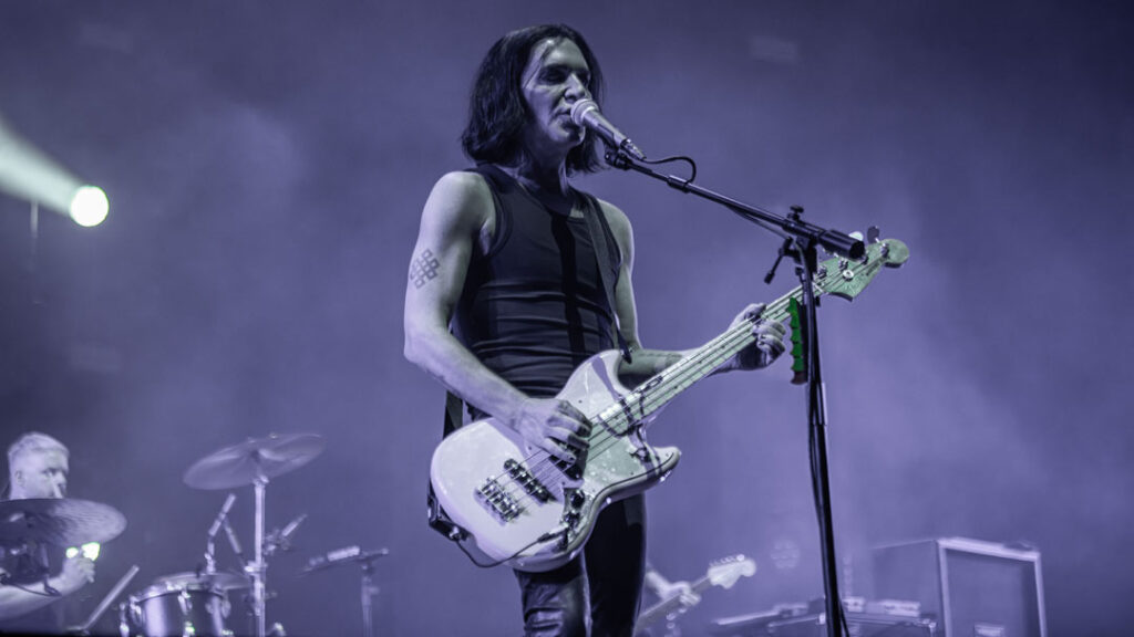 Der Sänger und Gitarrist Brian Molko steht in der Rockhal auf der Bühne und singt und spielt Gitarre. Molko ist ein dünner Mann mit scheitellangen schwarzen Haaren, er trägt ein dunkles, ärmelloses Oberteil und eine zerrissene Jeans. Im Hintergrund ist das Schlagzeug und ein Bass zu sehen.