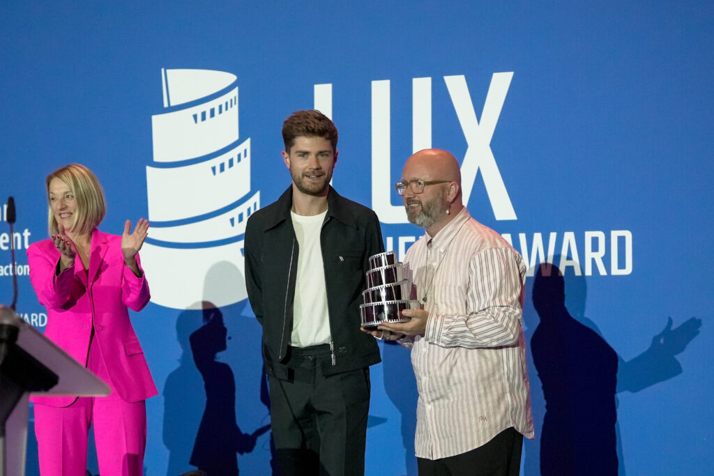 Lukas Dhont mit der applaudierenden Präsidentin des EU-Parlaments Roberta Metsola und einem Mann, der ihm den LUX-Filmpreis überreicht, auf der Bühne.