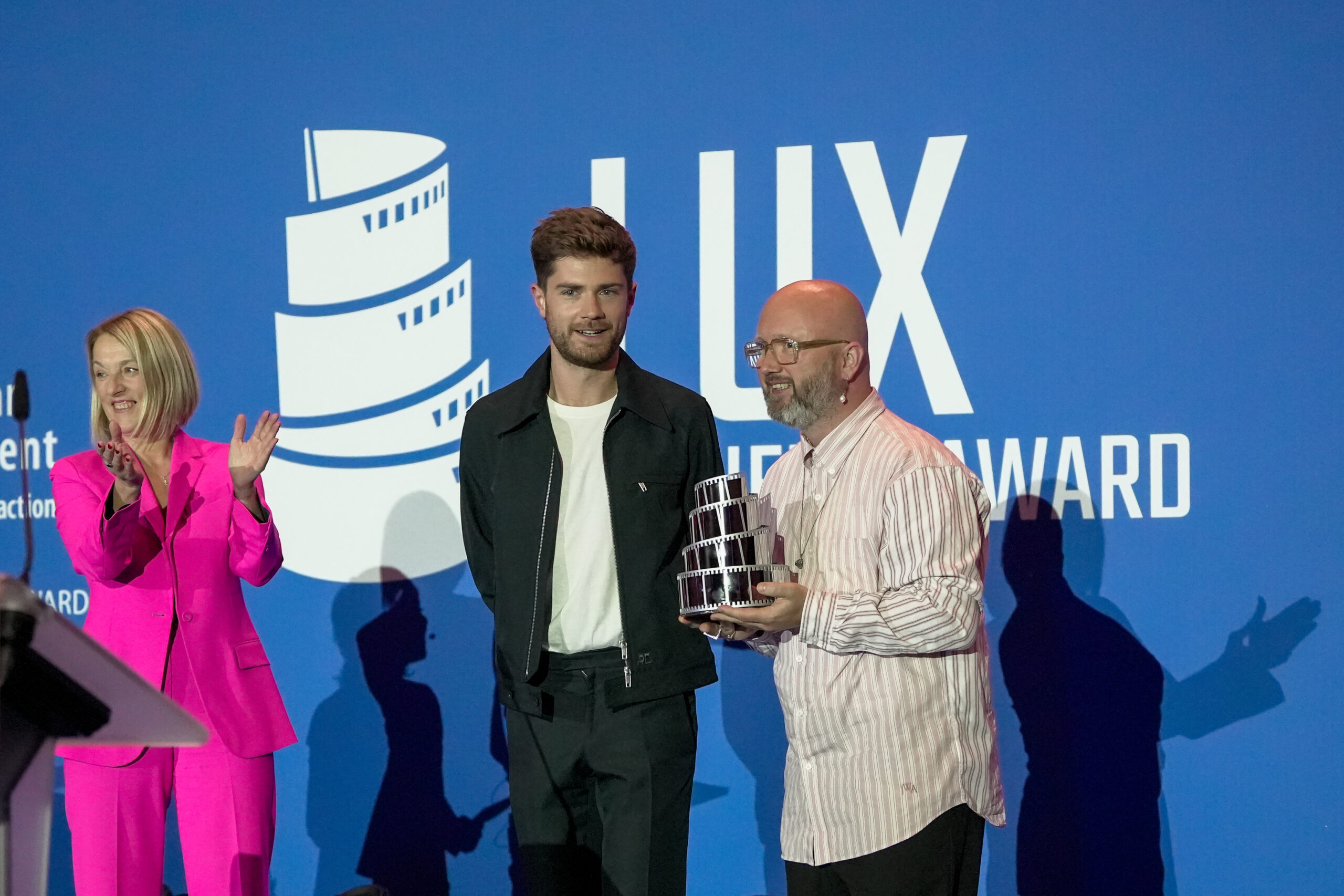 Lukas Dhont mit der applaudierenden Präsidentin des EU-Parlaments Roberta Metsola und einem Mann, der ihm den LUX-Filmpreis überreicht, auf der Bühne.