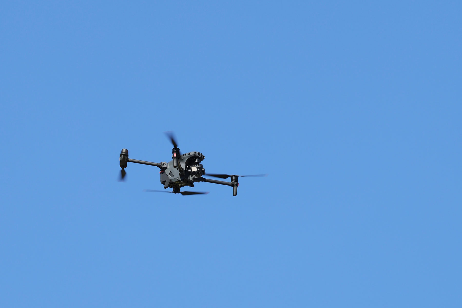 Eine Überwachungsdrohne vor blauem Himmel. Das Gerät hat einen hellgrauen Körper mit einer großen, gut sichtbaren Kamera und mehreren Linsen. Es fliegt durch vier Propeller, die an langen Armen an der Drohne befestigt sind.